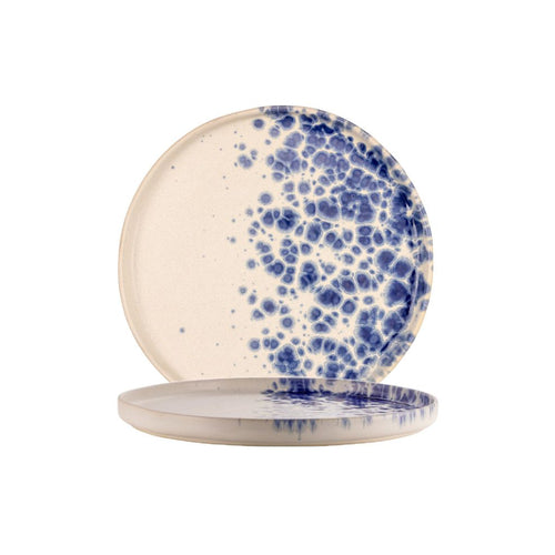 Due piatti rotondi in ceramica beige con decorazione macchie blu concentrata su metà superficie, sfondo bianco semplice.