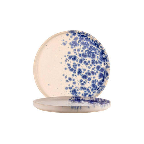 Due piatti rotondi in ceramica beige con macchie blu irregolari, uno appoggiato e uno in piedi sullo sfondo bianco.