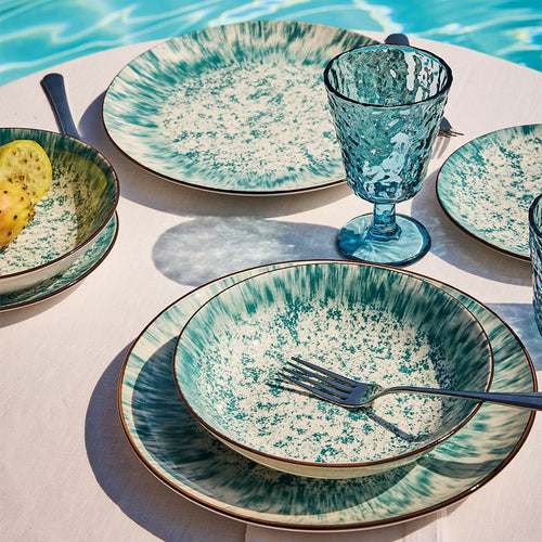 Set aus aquagrünen Keramik-Tellern und -Gläsern mit Spritz-Effekt, Metallbesteck, auf weißem Tisch nahe dem Pool.