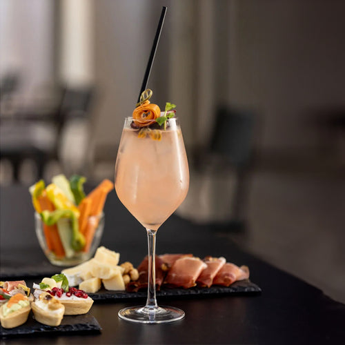 Transparentes Weinglas mit rosafarbenem Cocktail, dekoriert mit gerollter Orangenscheibe, grünem Blatt und Beeren, auf schwarzem Tisch mit Antipasti und