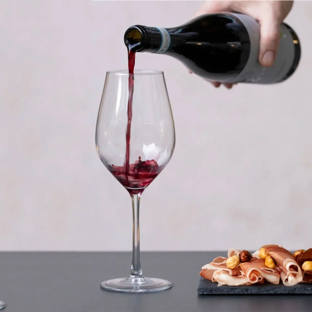 Vino tinto servido en copa de vidrio transparente con plato de pizarra con jamón crudo y avellanas sobre mesa negra.