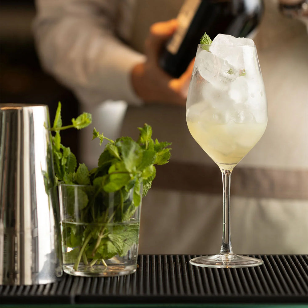 Copa de vino de vidrio transparente que contiene cóctel blanco con hielo y hoja de menta sobre tapete negro junto a copa con menta