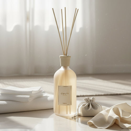 Diffusore d'ambiente in vetro opaco beige con bastoncini in legno, accanto a panni e sacchetto di tessuto bianco su superficie chiara.
