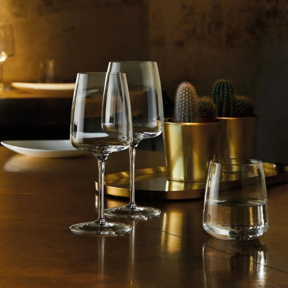 Dos vasos de vidrio transparente y un vaso bajo con agua sobre mesa marrón brillante, dos jarrones dorados con cactus al fondo.