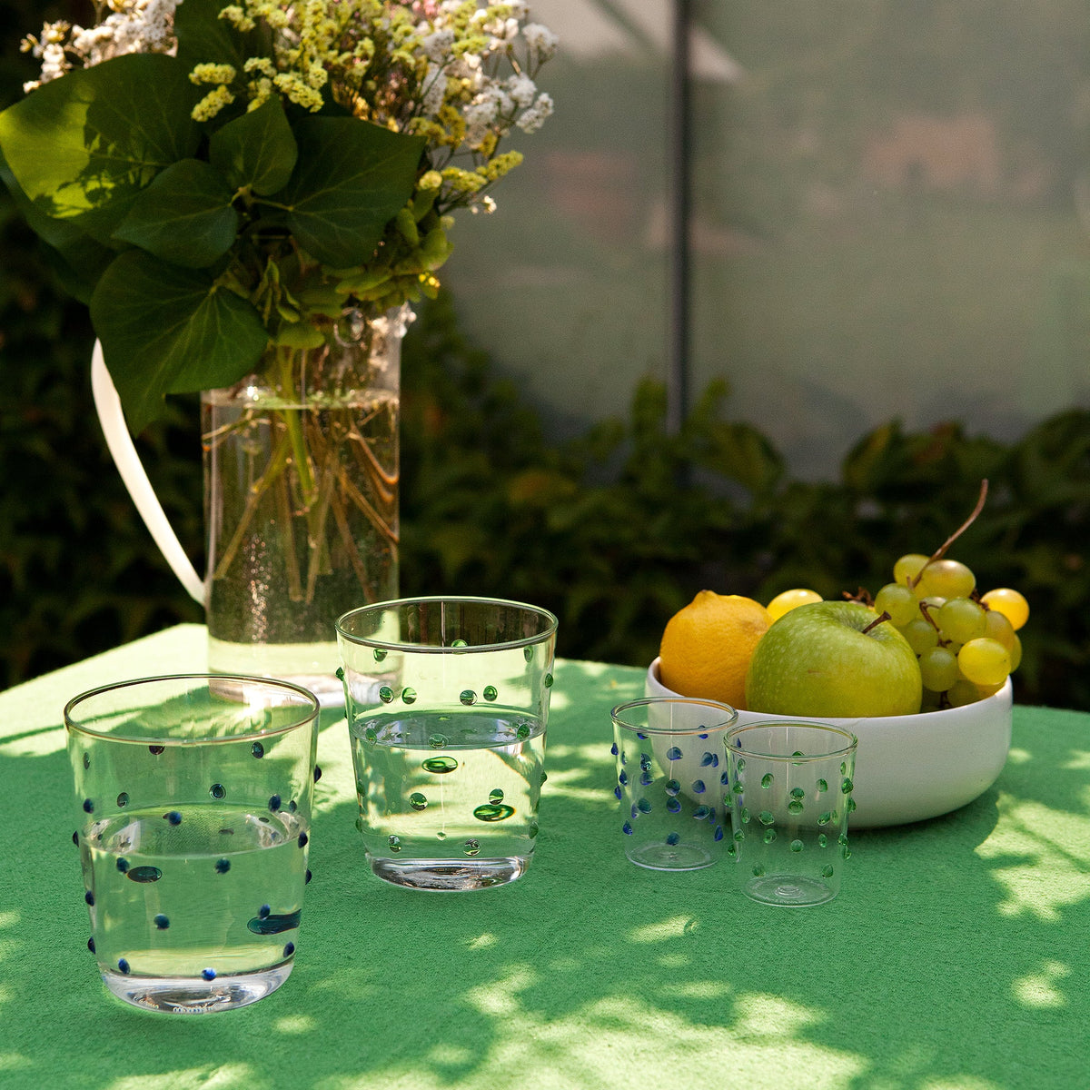 tavolo verde con due bicchieri in vetro trasparente con bolle colorate contenenti acqua, due bicchierini piccoli, vaso trasparente con fiori e
