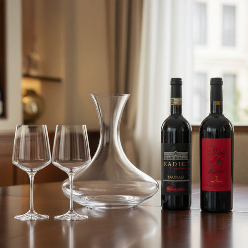 Due bottiglie di vino rosso con etichette scure, una caraffa in vetro trasparente e due calici di vetro su tavolo di legno.