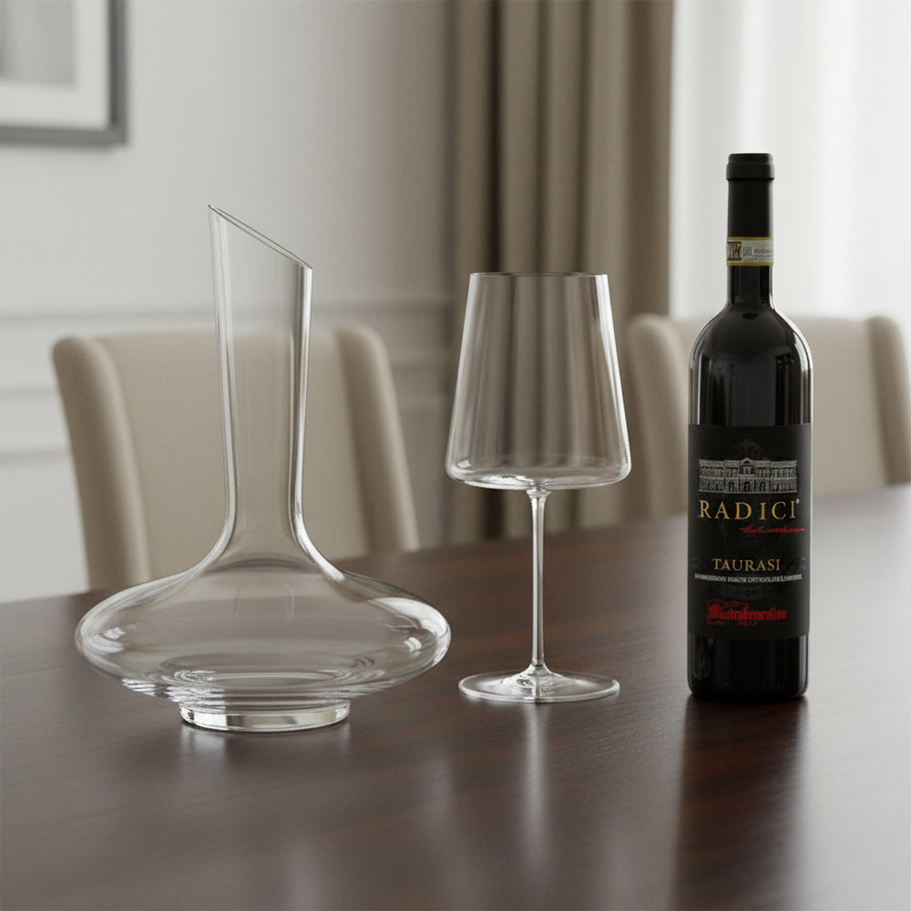 Caraffa di vetro trasparente, calice da vino con stelo sottile e bottiglia di vino rosso Radici Taurasi su tavolo in legno, sfondo soggiorno elegante.