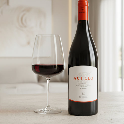 Achelo Rotweinflasche mit weißem Etikett und orangefarbener Schrift neben transparentem Weinglas mit eingegossenem Rotwein, auf Oberfläche
