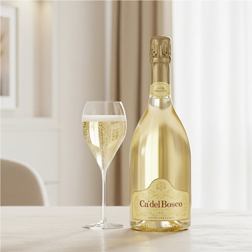 "Ca' del Bosco" Schaumweinflasche neben einem Glas mit Drink. Heller Tisch, beige Wände und Fenster mit hellen Vorhängen.