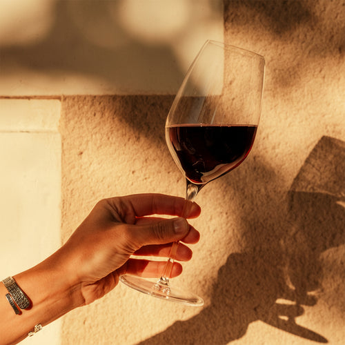 mano con bracciale regge calice di vetro trasparente con vino rosso contro parete beige illuminata da luce calda