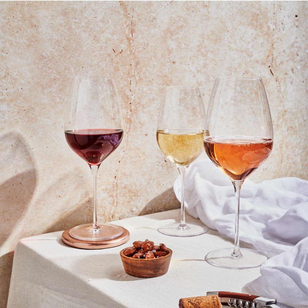 trois calices en verre transparent avec vin rouge, blanc et rosé, petit bol en bois avec amandes, nappe blanche et mur beige en arrière-plan