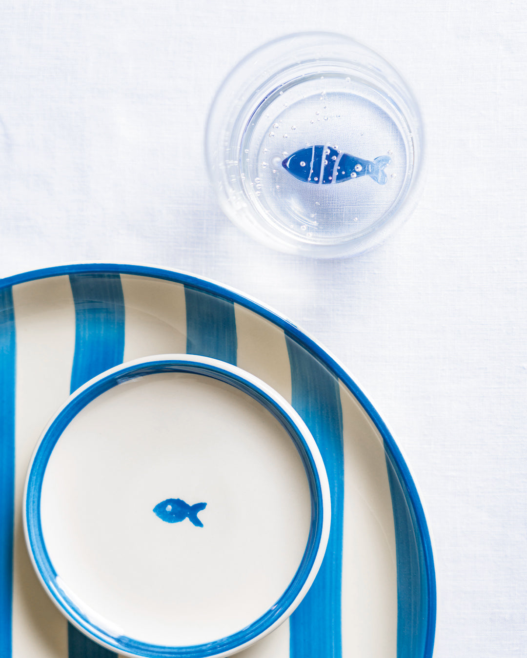 Witte keramische borden met strepen en blauwe vis, naast waterglas met decoratieve blauwe vis en bellen, op wit tafelkleed.