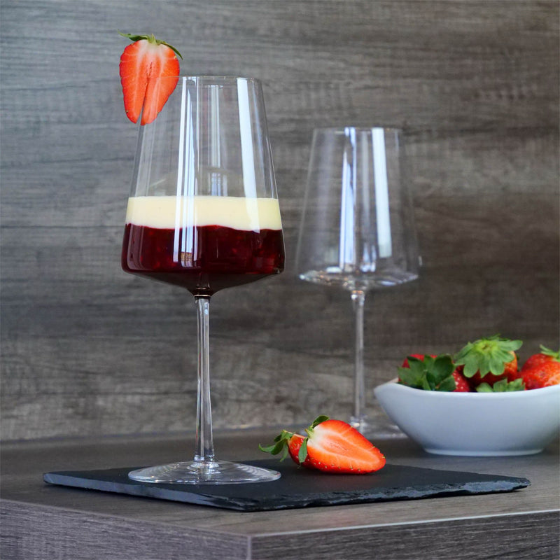 Calice di vetro trasparente con dessert a strati rosso e crema, fragola a metà sul bordo, altra fragola e ciotola con fragole sul piano scuro in legno