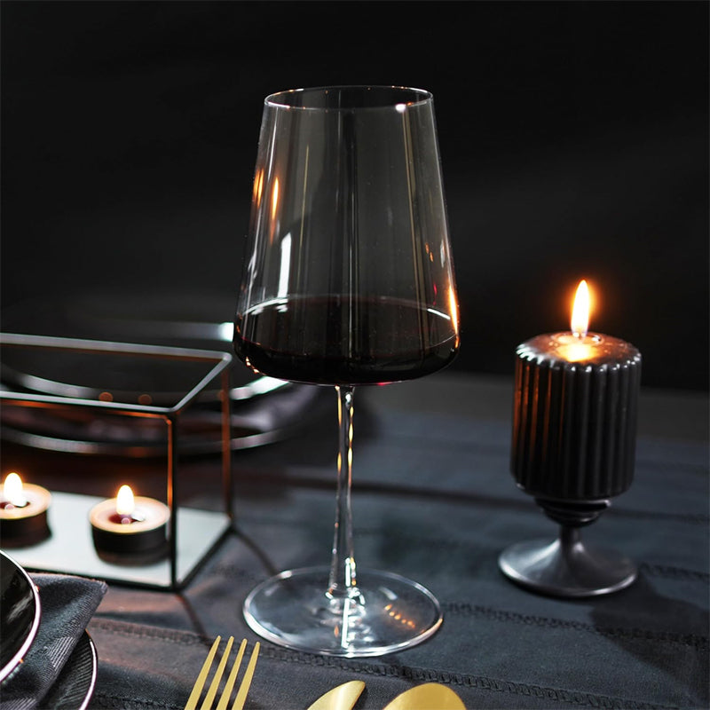 Calice di vetro trasparente con vino rosso su tavola scura, accanto candela nera accesa e porta tea light nero con tea light accese.