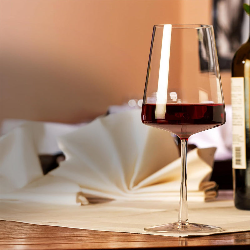 Calice di vetro trasparente con vino rosso, su tavolo di legno chiaro con tovagliolo bianco piegato e bottiglia verde sullo sfondo sfocato