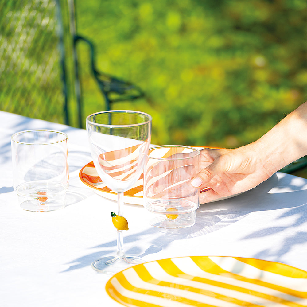 Bicchieri trasparenti in vetro con decorazione limone su tavolo bianco con piatti gialli e bianchi a righe, all'aperto su sfondo verde sfocato.