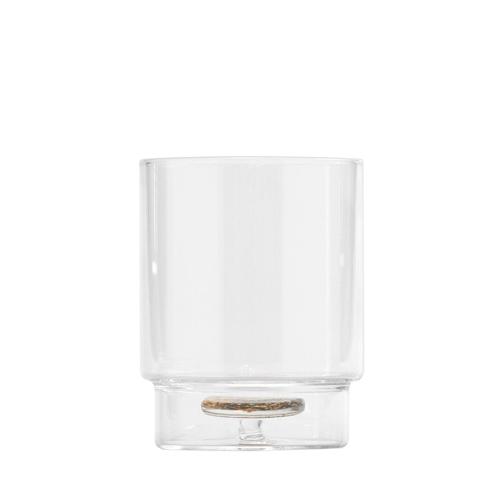 Bicchiere trasparente in vetro con base piatta e elemento circolare decorativo marrone e nero sul fondo, sfondo bianco.