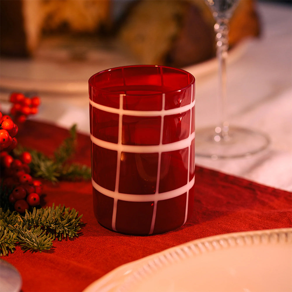 Verre cylindrique rouge avec motifs quadrillés blancs sur nappe rouge, décorations de Noël et assiettes blanches en céramique.