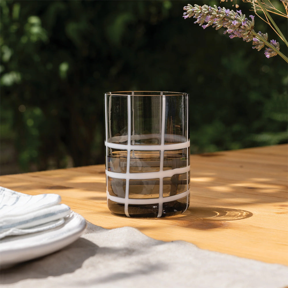 Verre transparent avec motif à carreaux blancs sur table en bois clair en extérieur avec assiette et serviette sur fond vert