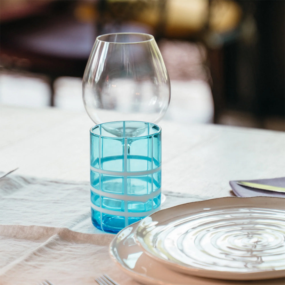 Verre transparent bleu clair avec motif quadrillé blanc, posé à côté d'un calice transparent sur table dressée.