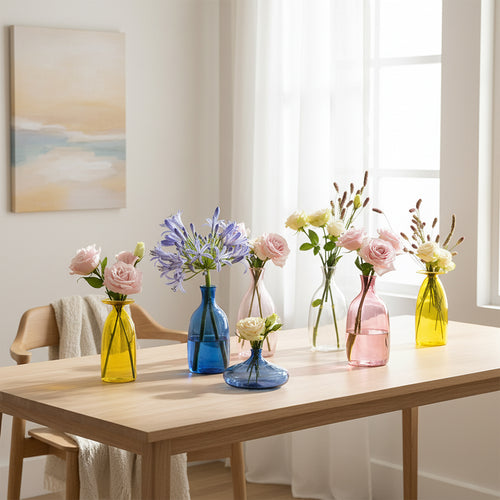 Set di sette vasi di vetro colorati (giallo, blu, rosa, trasparente) con fiori freschi su tavolo di legno chiaro in ambiente illuminato da finestra.