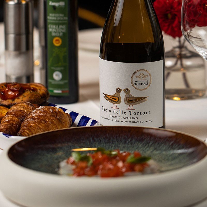 Bottiglia di vino bianco Bacio delle Tortore con etichetta beige e disegno di due tortore, su tavola con croissant, piatto e bottiglia sullo sfondo.