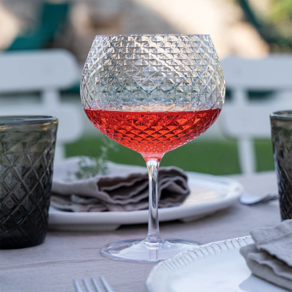 Verre à vin en verre transparent avec motif gravé contenant une boisson rouge, sur table dressée en plein air avec assiettes et serviettes grises