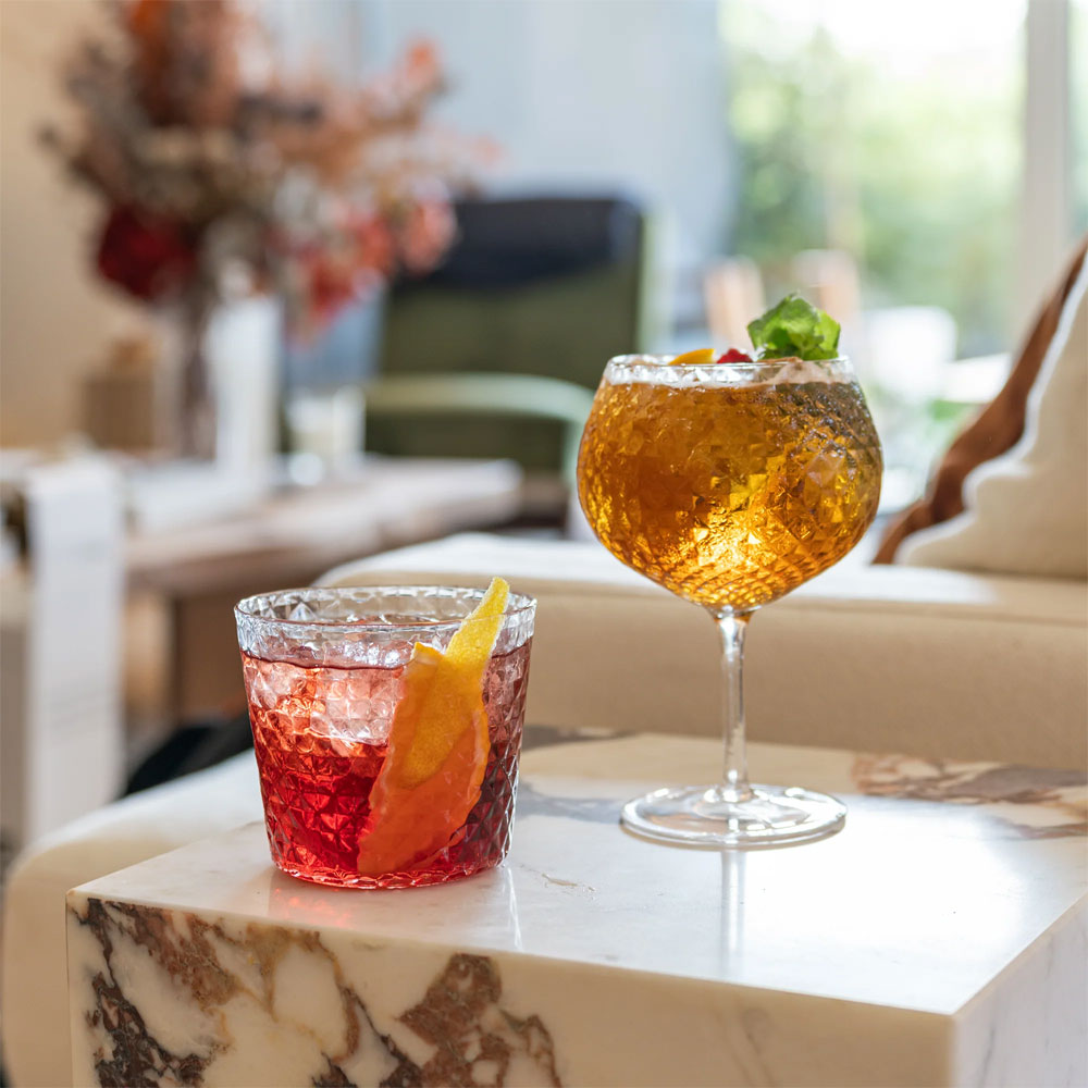 deux cocktails en verres facettés, un rouge avec zeste d'agrumes et un ambré avec feuille de menthe, sur table en marbre blanc