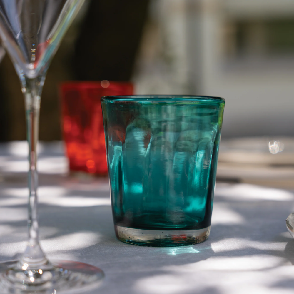 Dik turquoise glas met transparante bodem op wit tafellaken, rood glas en transparant wijnglas op de achtergrond.