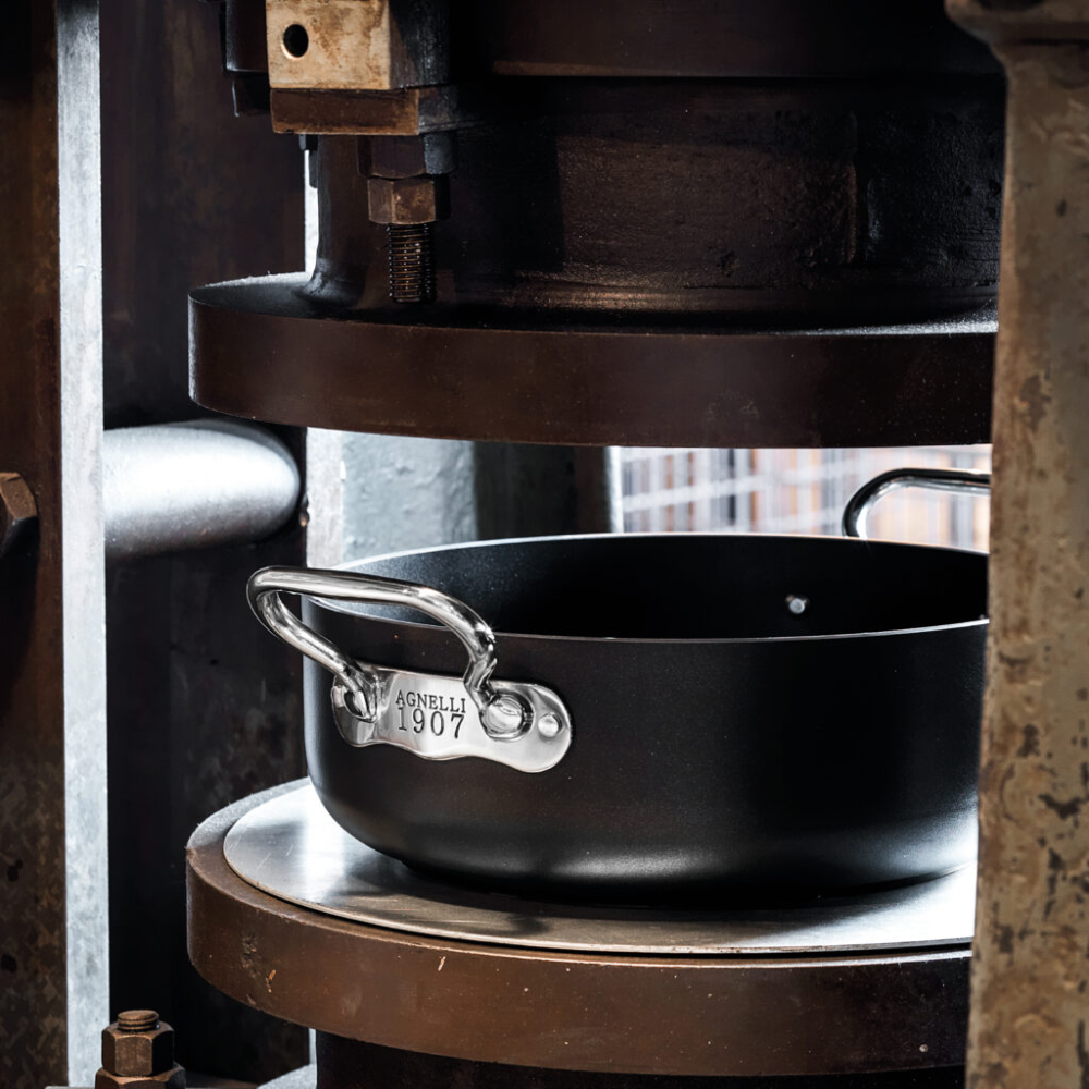 Pentola nera con manici in metallo lucido e targhetta "AGNELLI 1907" inserita in una pressa industriale di metallo scuro.