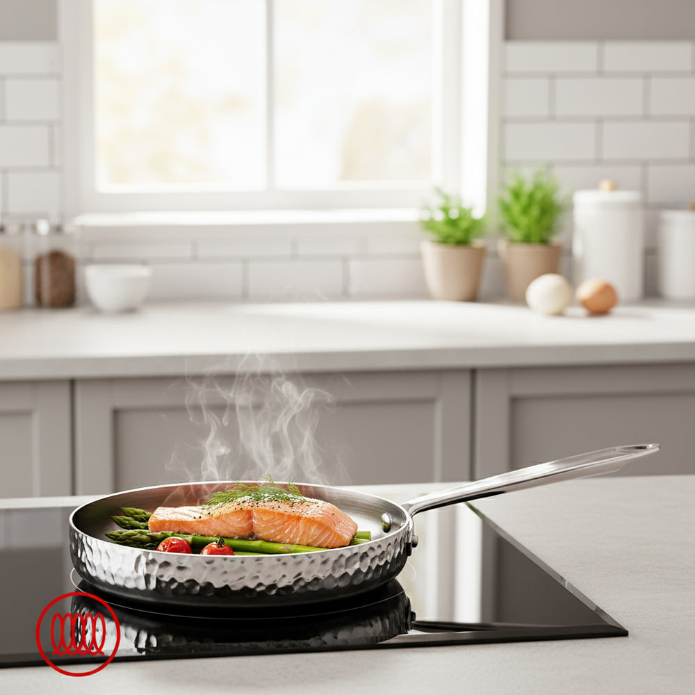 Padella martellata in acciaio inox con salmone rosa, asparagi verdi e pomodori ciliegia, su piano cottura nero in cucina luminosa.