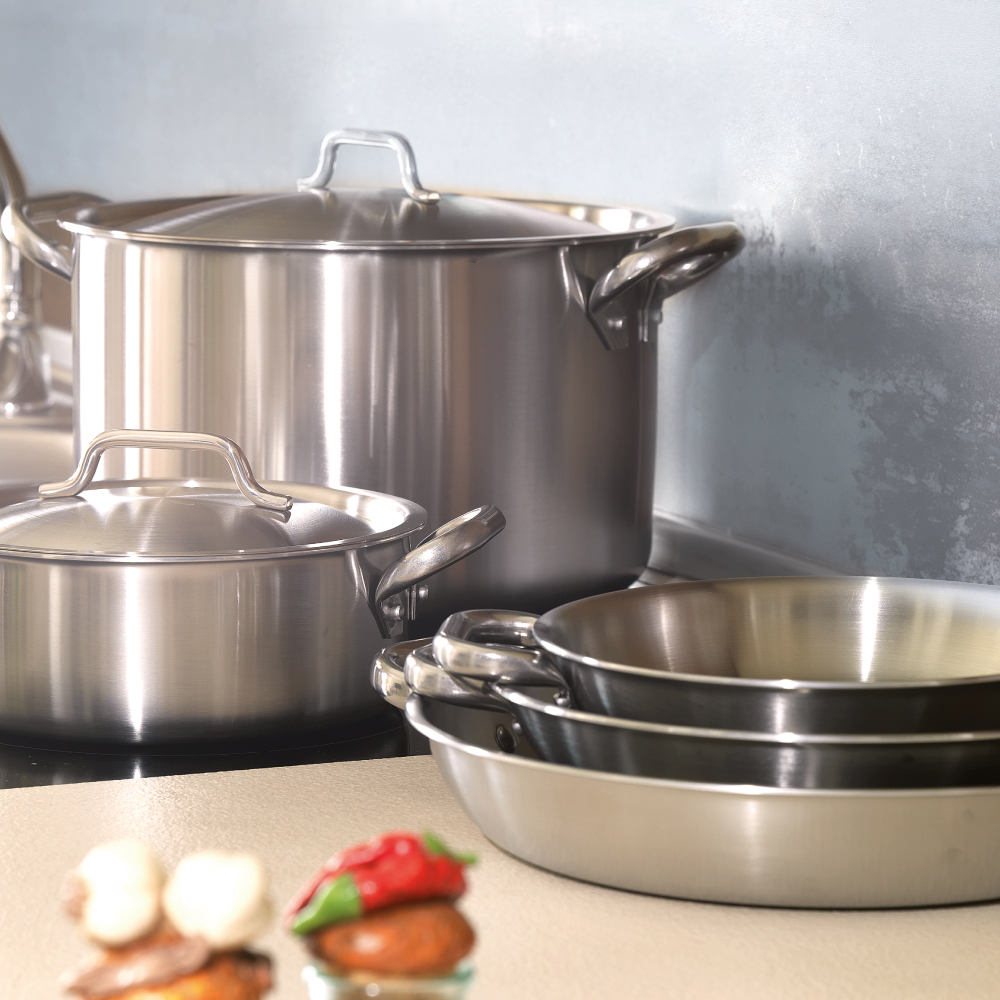 Set di pentole e padelle in acciaio inox lucido con coperchi e manici, collocate su piano cucina beige con sfondo grigio.