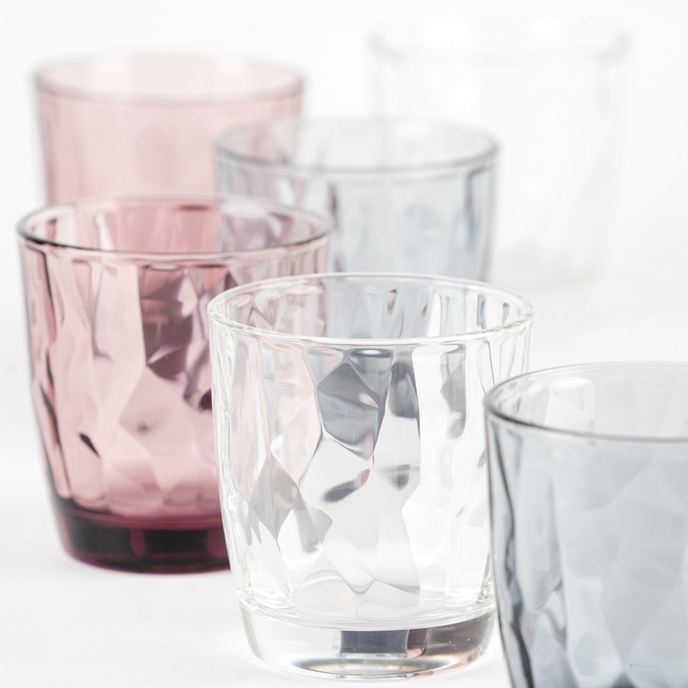 Sette bicchieri trasparenti di vari colori (rosa, grigio, chiaro e trasparente) con superficie sfaccettata.