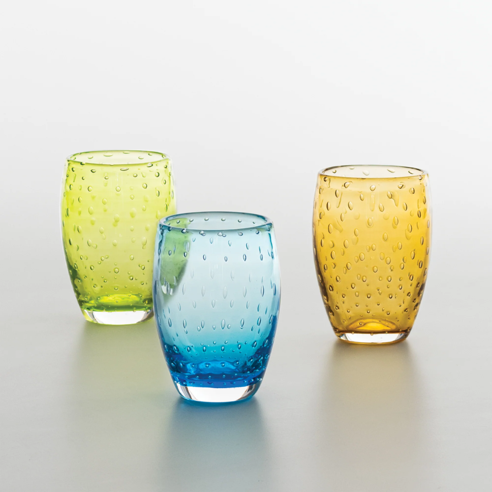 Tre bicchieri di vetro trasparente puntinati, nei colori verde, azzurro e arancione, su superficie chiara uniforme.