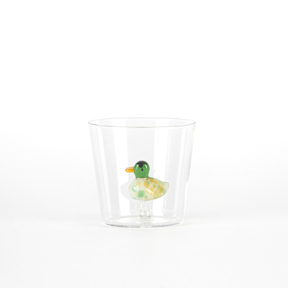 Un vaso transparente contiene una figura de pato de colores, con cuerpo verde y manchas amarillas. Fondo blanco.