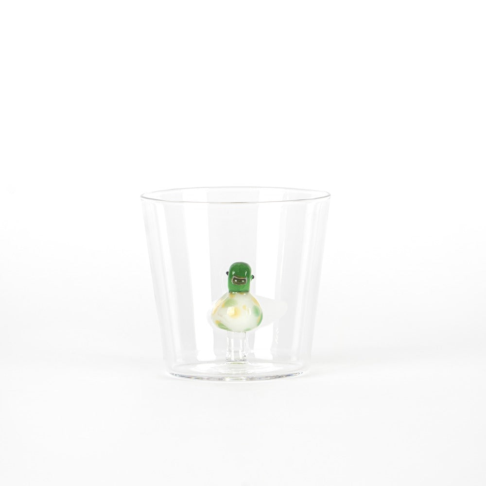Figurita verde con una sonrisa dentro de un vaso de vidrio transparente, sobre fondo blanco.