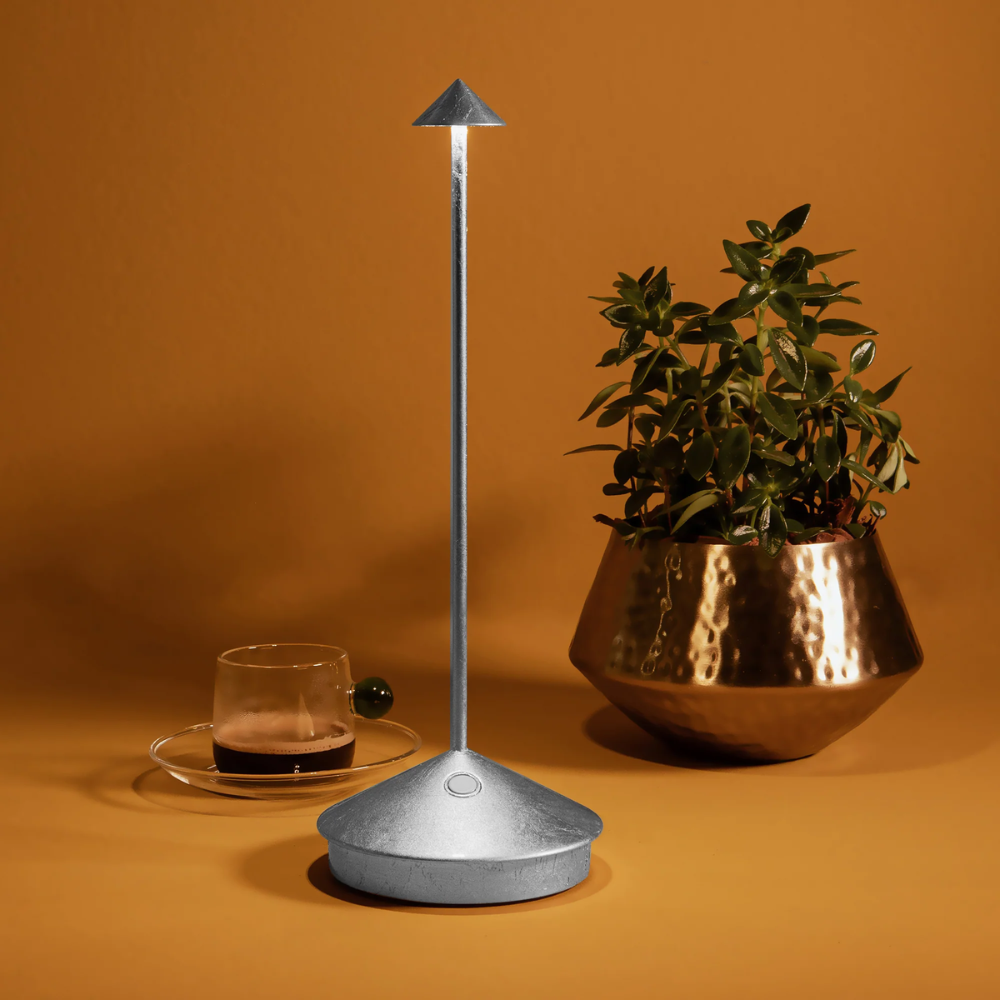 Lampada da tavolo argento in metallo con base tonda e paralume a forma di cono, accanto a tazza di caffè e vaso con pianta verde su sfondo