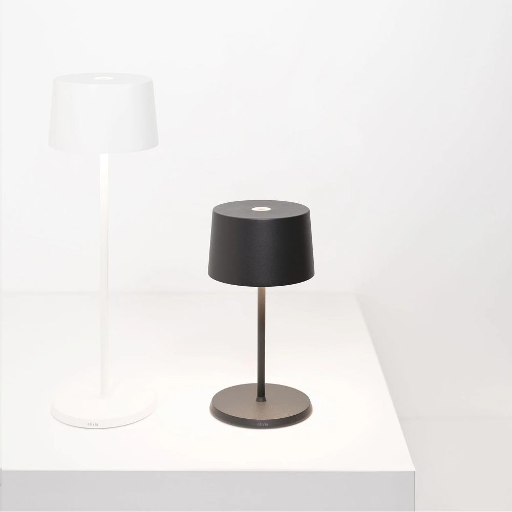 lampada da tavolo nera con paralume cilindrico in metallo opaco e base rotonda, posizionata su superficie bianca vicino a paralume bianco