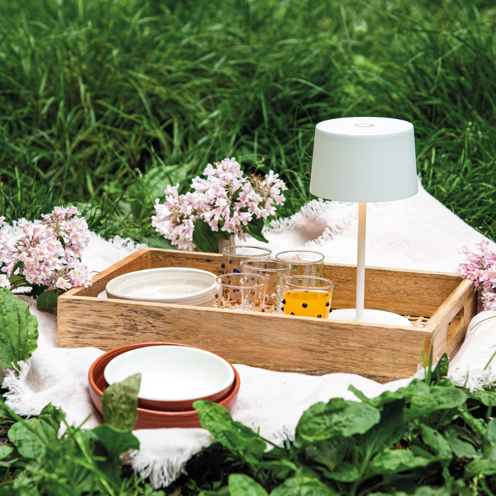 Plateau rectangulaire en bois avec lampe blanche, quatre verres décorés, assiettes blanches et fleurs roses sur nappe blanche, pelouse verte.