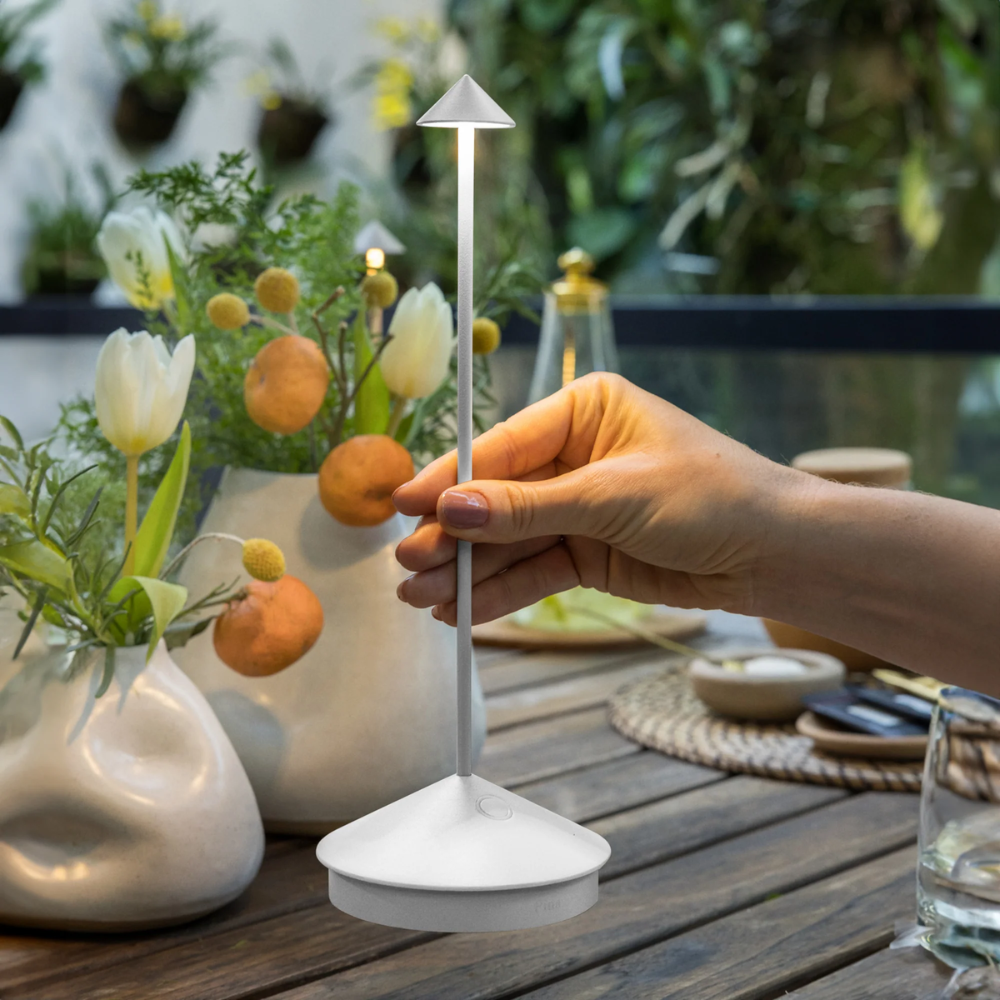 Lampada da tavolo bianca a forma di fungo con base triangolare tonda, in un ambiente esterno con fiori e frutti decorativi.