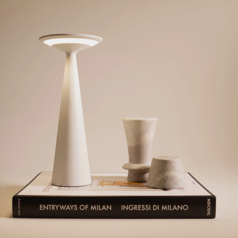 Weiße konische Lampe mit rundem Diffusor, zwei kleine Vasen aus hellem Marmor und Designbuch mit weiß-schwarzem Cover.
