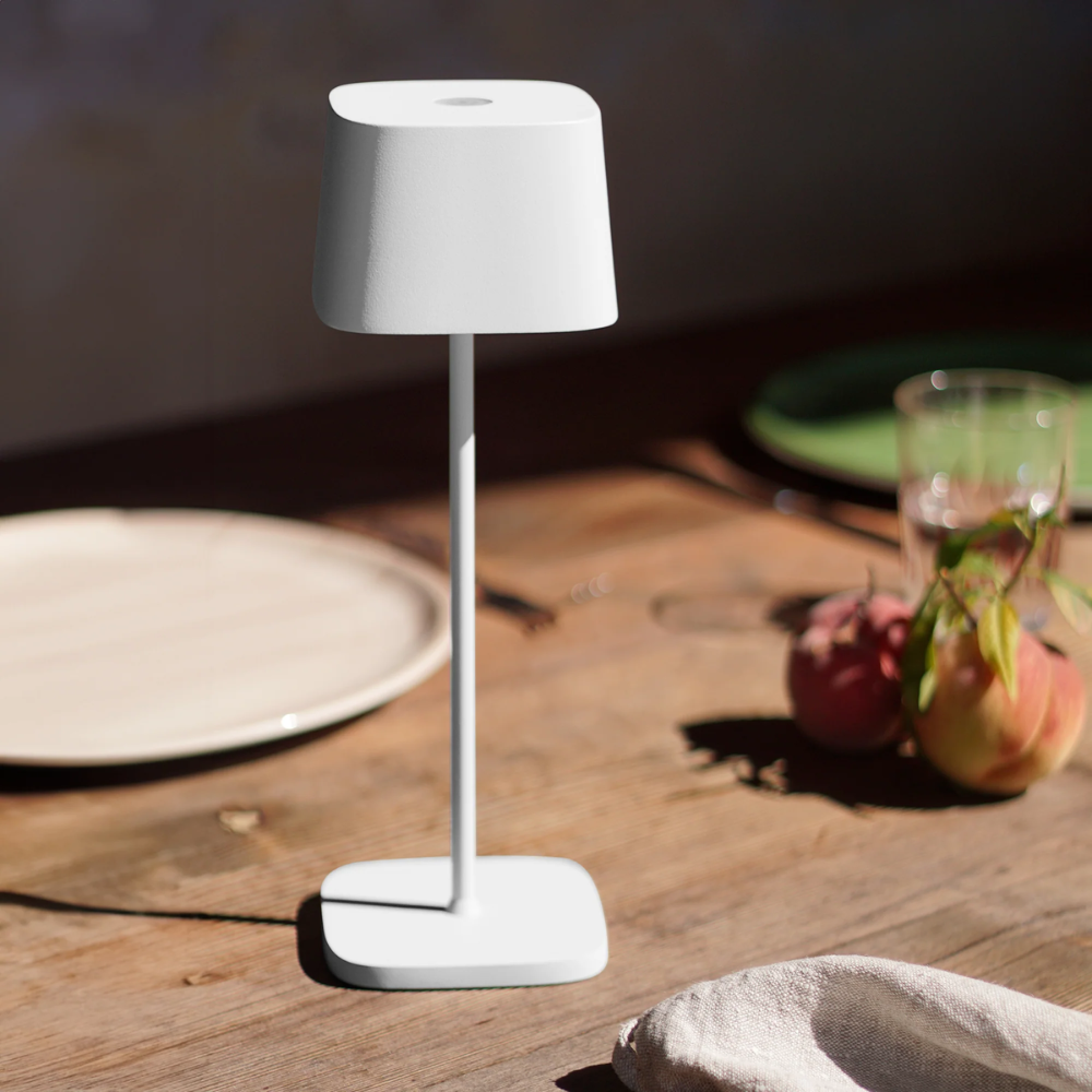 Weiße Lampe mit minimalistischem Design, quadratischer Basis und Schirm auf Holztisch mit Tellern, Glas und Obst.