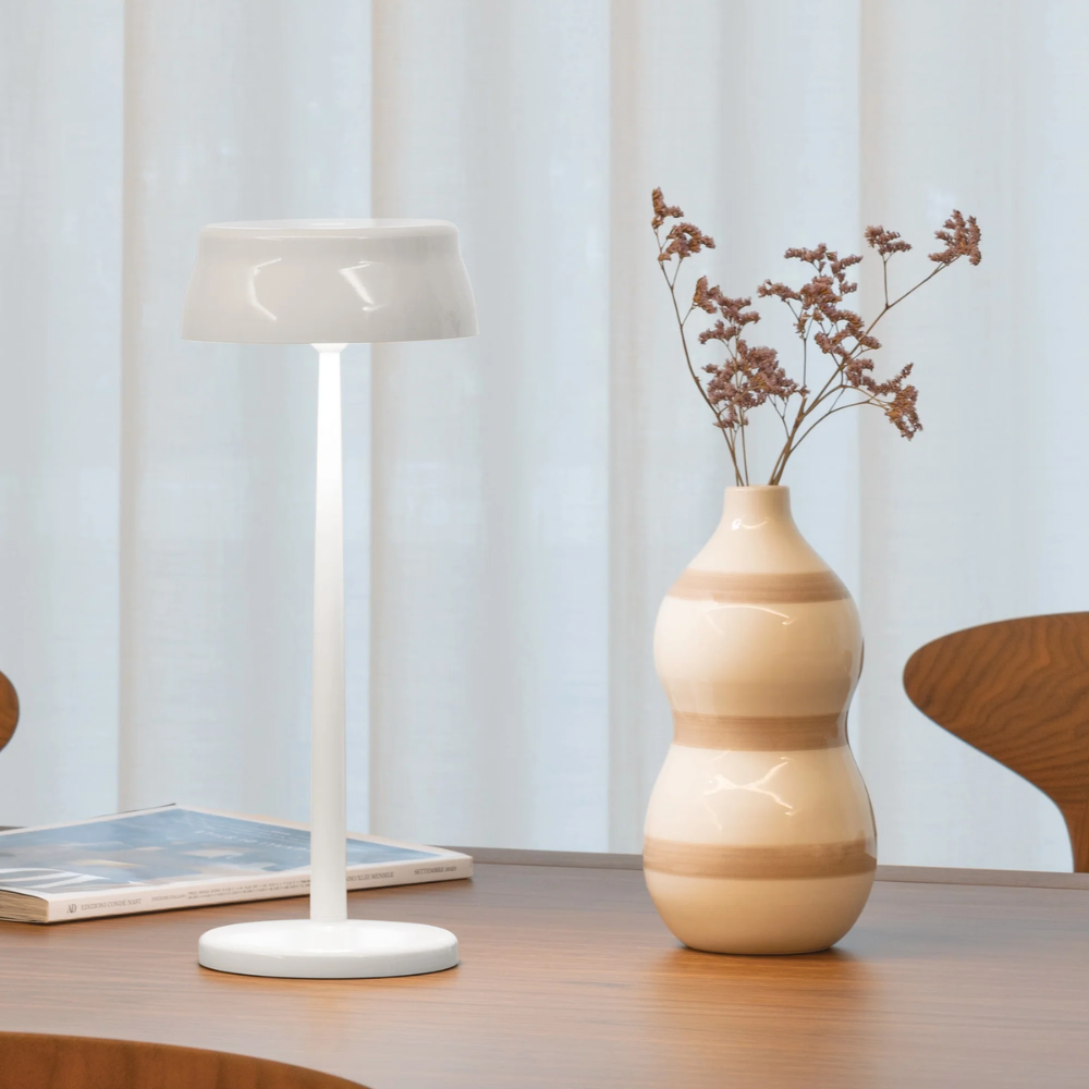 Lampe de table blanche en métal avec abat-jour rond, vase en céramique beige et marron avec fleurs séchées sur table en bois.