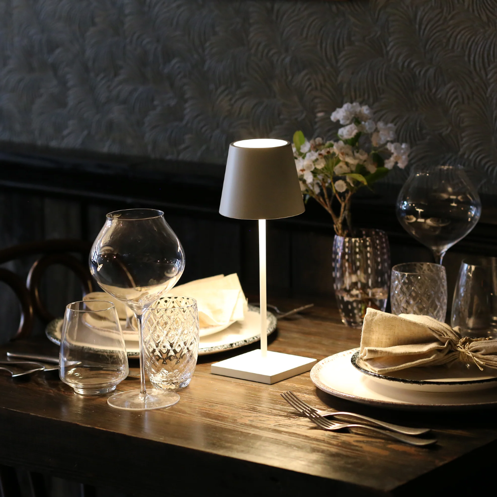 Lampada da tavolo bianca a forma di abat-jour minimalista accesa su tavolo in legno apparecchiato con vetri, piatti e fiori bianchi.