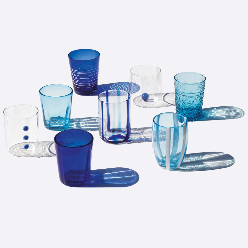 Set di otto bicchieri di vetro in varie tonalità di blu, trasparenti e opachi, con motivi a righe e decorazioni, su sfondo chiaro.
