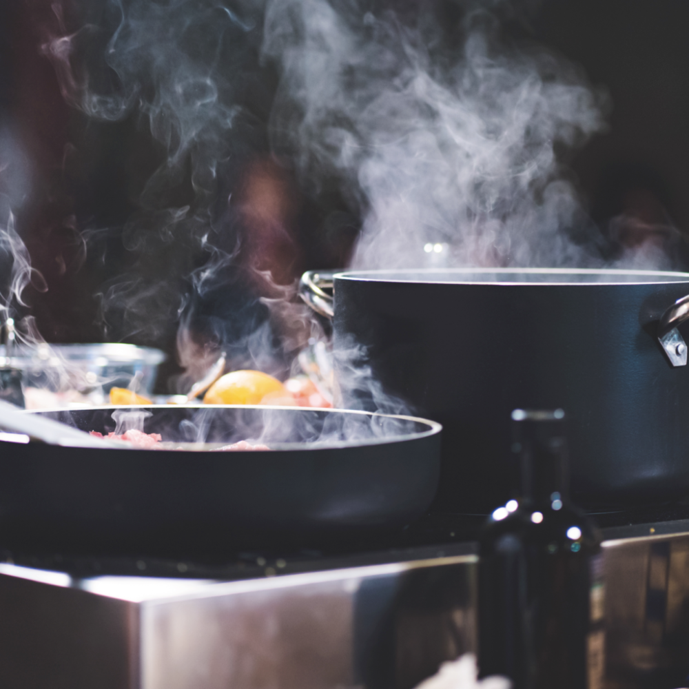 Schwarzer Topf und Pfanne mit Metallgriffen, beide dampfend auf dem Herd in einer modernen Küche.