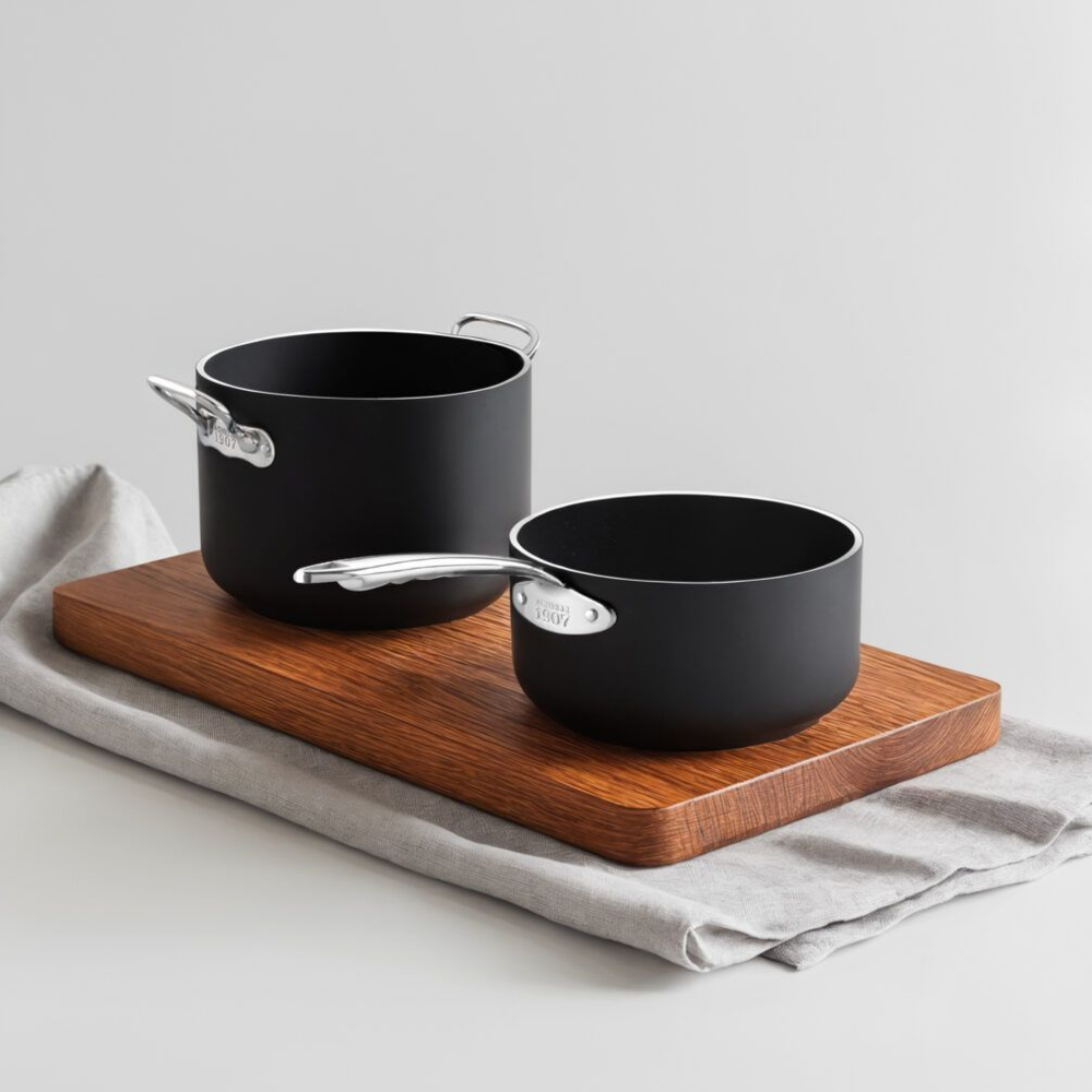 Twee zwarte pannen met glanzende metalen handvatten, een met dubbel handvat en een met lang handvat, op houten snijplank en grijze stof.
