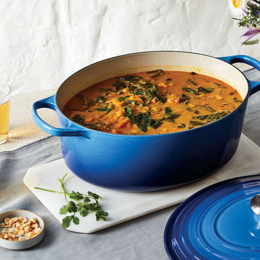 Cocotte ovale bleue avec intérieur blanc contenant soupe chaude aux légumes verts, sur planche blanche avec assiette de noisettes et verre