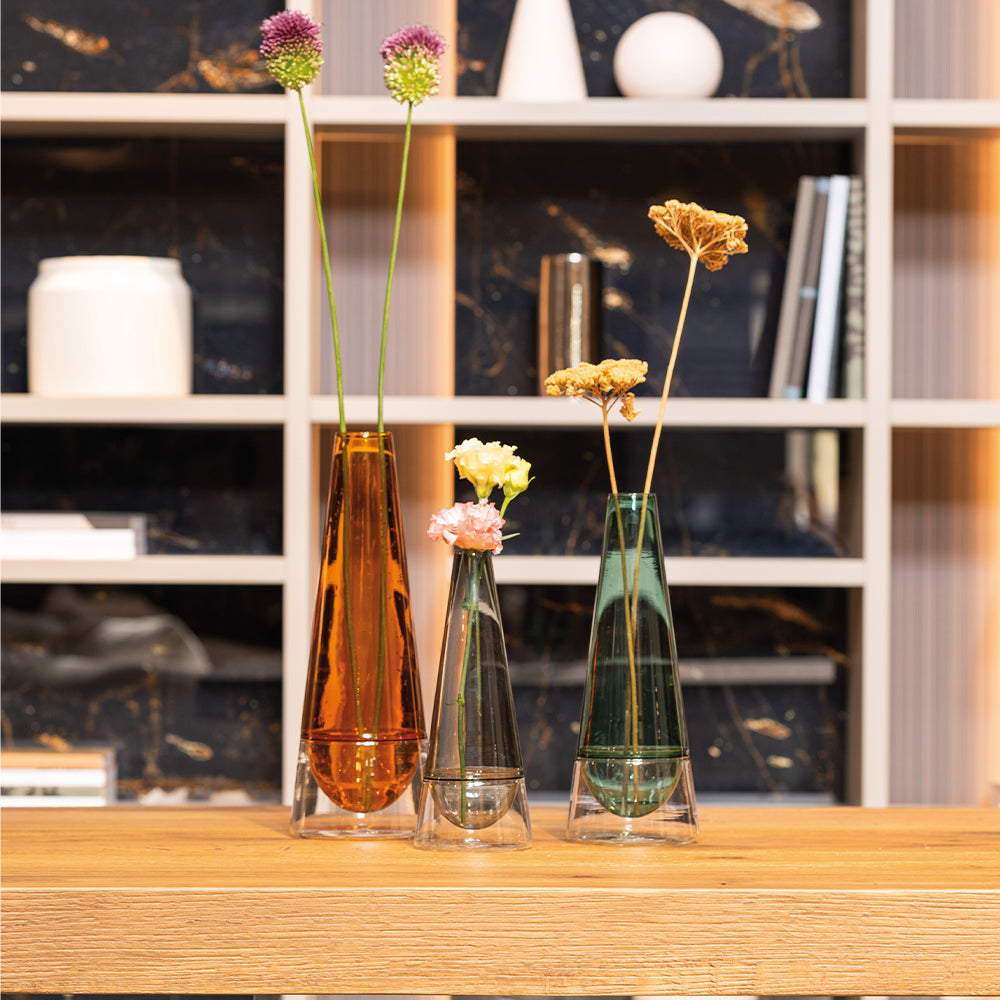 Tres jarrones de vidrio de distintas alturas con flores, sobre mesa de madera delante de una estantería moderna.