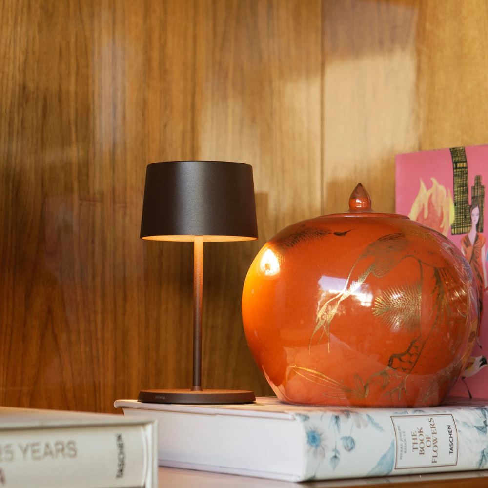 Lampe de table avec base et abat-jour noir mat à côté d'un vase orange décoré d'un motif doré sur surface en bois avec livres et
